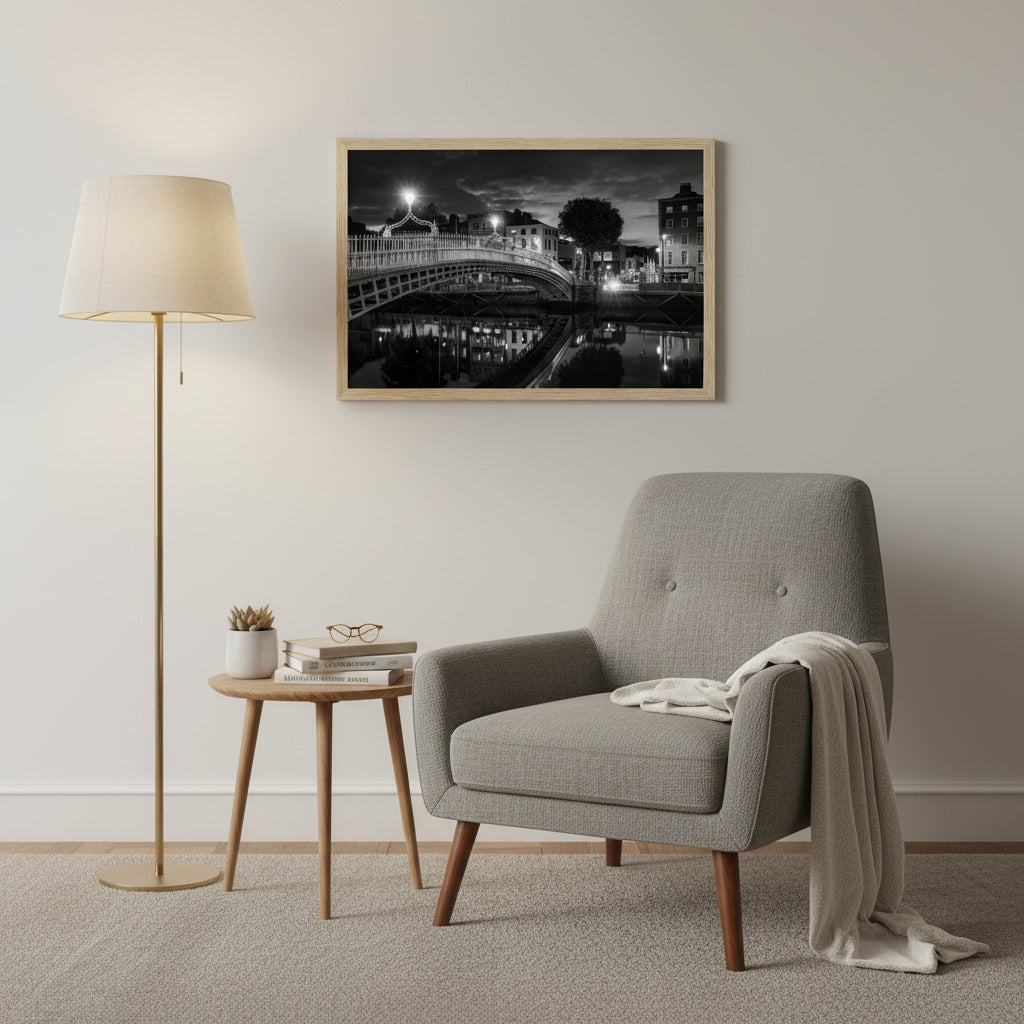Black and white night view of Ha’penny Bridge over the River Liffey in Dublin, framed in a natural wood frame, 30x45 cm.Cosy reading nook with mid-century armchair and floor lamp, 30x45 cm Ha’penny Bridge black and white photograph in natural wood frame on light wall, stack of books and plant, warm evening lighting, realistic framed print